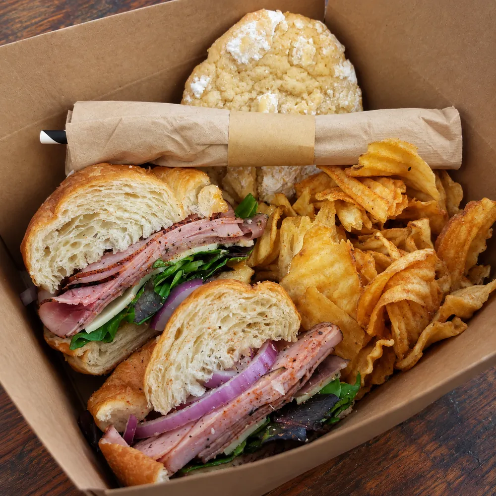 An open kraft take-out box: a croissant sandwich of sliced ham, cheese, red onion and greens cut in half, a pile of house-fried chips, a sugar cookie and napkin-wrapped cutlery