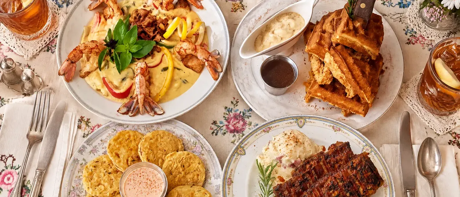 Plates from the lunch and supper menu seen from above: shrimp and grits, chicken and waffles with cream gravy, fried green tomatoes with remoulade and glazed meatloaf, with glasses of sweet tea