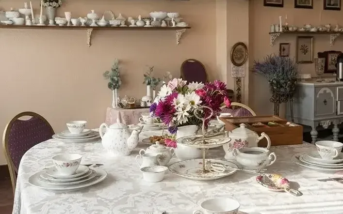 The Magnolia Room set for a tea: a round table in white damask with rose-patterned china, teapots and a tiered stand around a bouquet of dahlias and daisies, purple and gold chairs, and milk-glass on the shelves behind