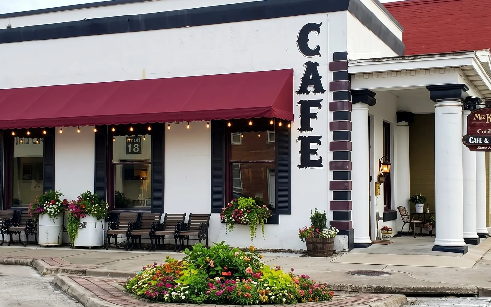 The Cotillion on Main Street: a white building with CAFE painted down the corner in tall black letters, a burgundy awning hung with string lights over shuttered windows, benches and flower planters along the sidewalk, and a columned porch at the front door