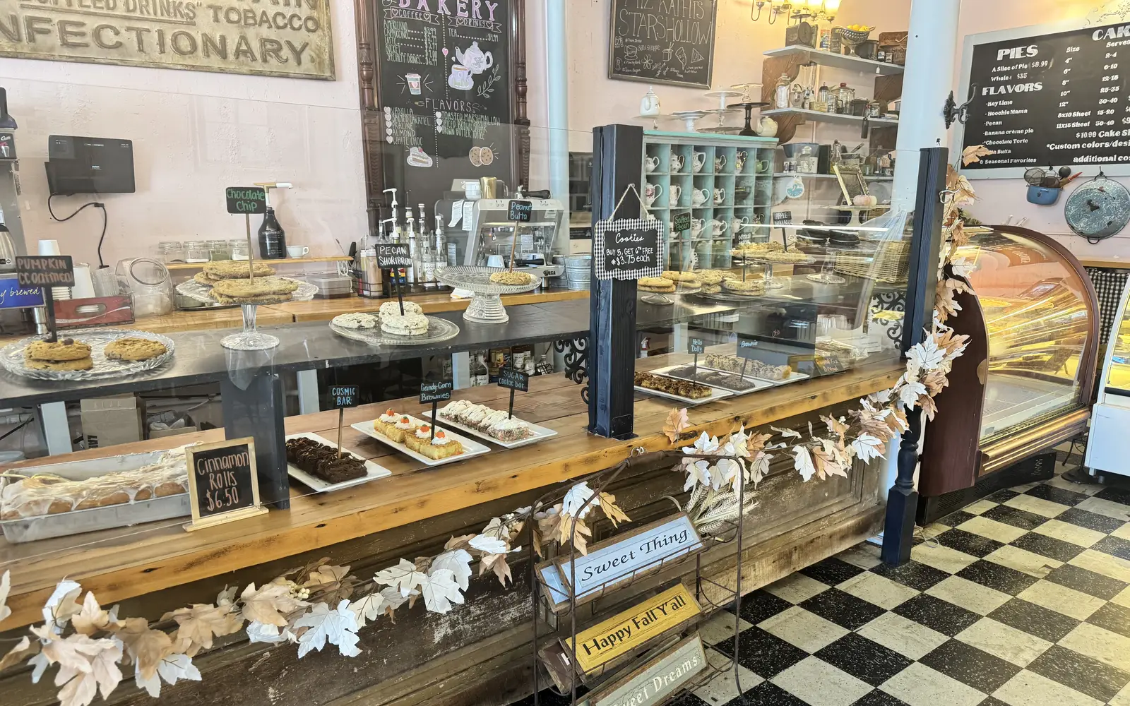The Sweetery counter: a long rustic wooden counter of cookies, brownies and dessert bars under glass, a lit pastry case at the end, chalkboard menus on pink walls and a black-and-white checkered floor