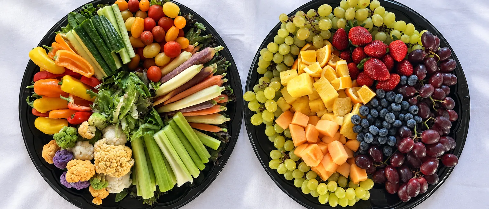 The Garden Vegetable Platter and the Fresh Fruit Platter side by side on white linen, seen from above