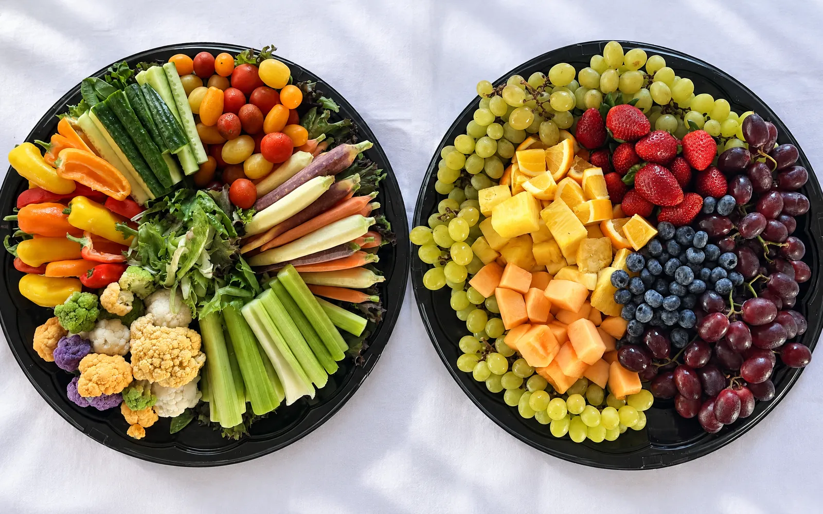 Two round catering platters side by side on white linen: the Garden Vegetable Platter of celery, cucumber, rainbow carrots, sweet peppers, cherry tomatoes and colored cauliflower, and the Fresh Fruit Platter of green and red grapes, strawberries, blueberries, pineapple, cantaloupe and orange wedges
