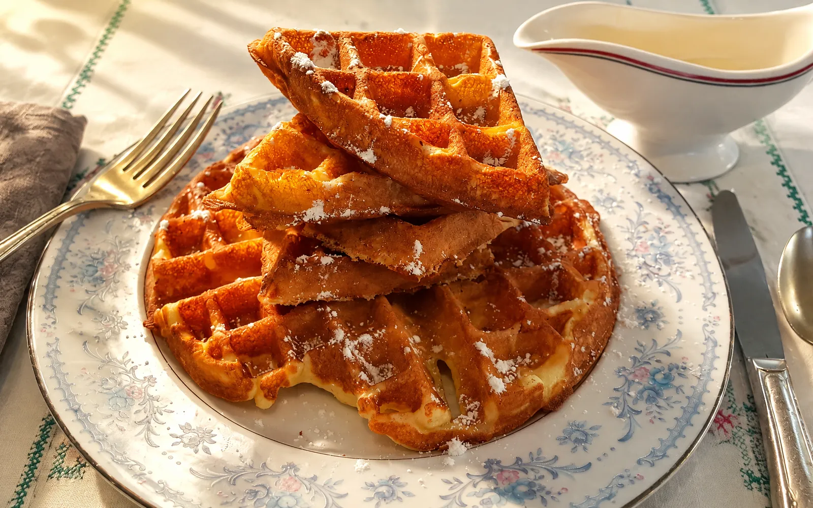 Golden waffles dusted with powdered sugar, stacked on vintage blue-and-white floral china beside a white gravy boat, on an embroidered tablecloth in morning sun